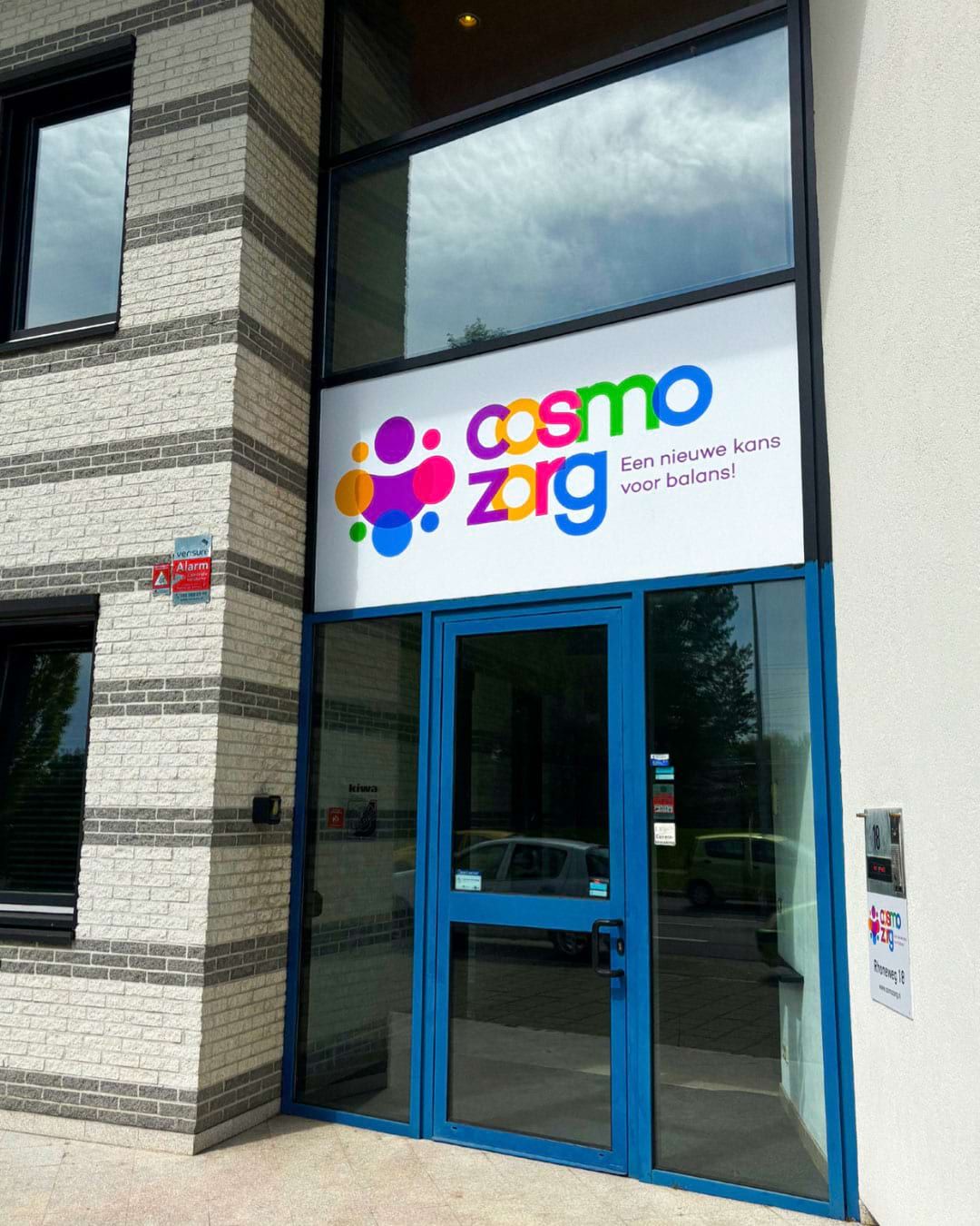 De entree van Cosmo Zorg aan de Rhôneweg 18, met blauwe kozijnen en een verlicht logopaneel naast de deur.